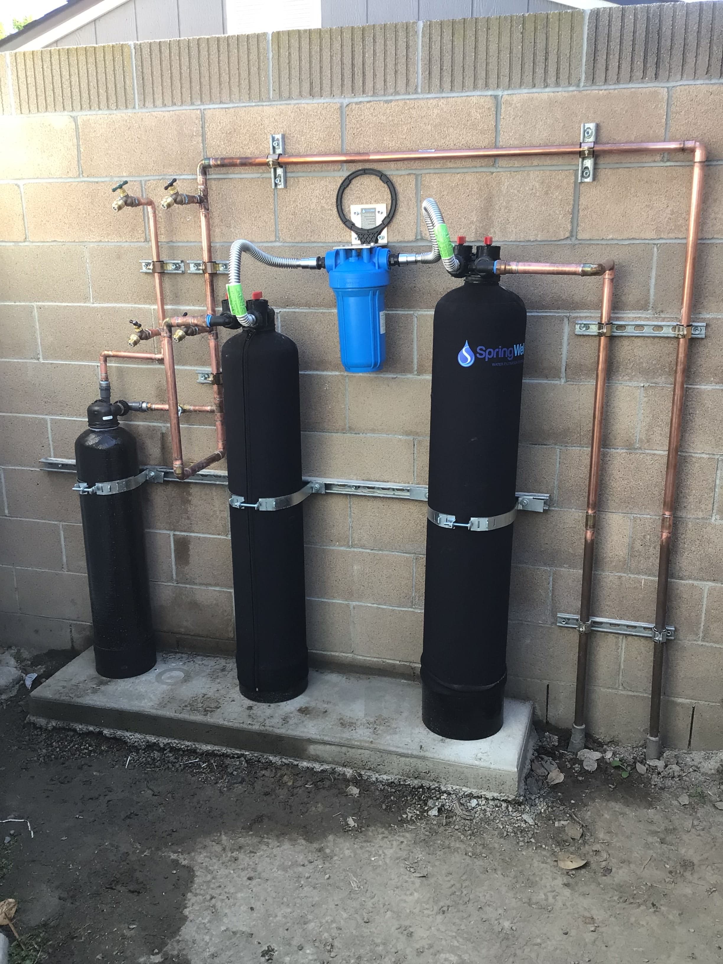 Whole-home water filtration system with sediment prefilter and twin media tanks, copper bypass manifold, and strapped supports on a CMU wall.
